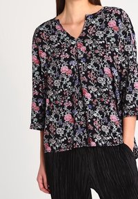 Sort blomsterbluse med v-hals, trekvartærmer og et mønster af lyserøde og blå blomster på en mørk baggrund.