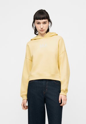 Young woman wearing a light yellow GAP hoodie and dark blue jeans, standing against a plain light gray background.