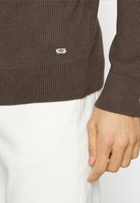 Pull marron à côtes avec des manches longues et une petite étiquette logo à l'ourlet, associé à un pantalon blanc. Texture douce et design ajusté.