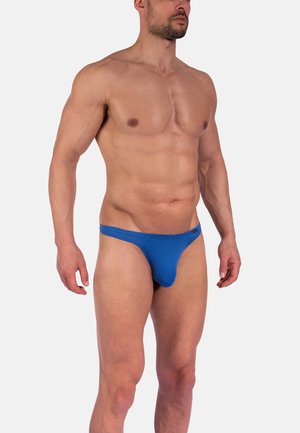 Svalnatý muž ve modrých pruhovaných bikinových kalhotkách, stojící s uvolněnýma rukama na jednoduchém bílém pozadí.