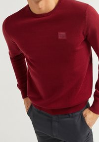 Burgundowy sweter dzianinowy z okrągłym dekoltem i ściągaczami, z małą naszywką z logo na piersi, zestawiony z ciemnymi spodniami.