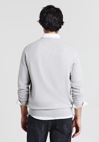 Mann mit dunklen Haaren, der einen hellgrauen Pullover über einem weißen Hemd und einer dunklen Jeans trägt, stehend, mit dem Rücken zur Kamera vor einem schlichten Hintergrund.