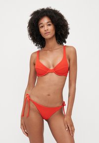Červený bikiny set s textúrovanou tkaninou, štruktúrovaným braletovým topom s nastaviteľnými ramienkami a nízko zvýšenými nohavičkami so šnurovaním po stranách.