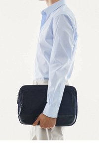Personne portant une chemise bleu clair et un pantalon beige tenant un sac d'ordinateur portable noir zippé à côté d'eux sur un fond blanc.