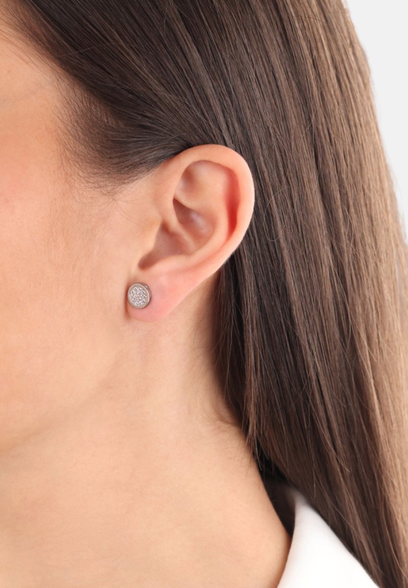 Pendiente de plata redondo con un patrón texturizado, montado sobre una base ligeramente rosa, mostrado contra un cabello liso y un fondo neutro.