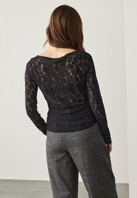 Haut à manches longues en dentelle noire avec un motif floral et un large décolleté, superposé sur une base ajustée, associé à un pantalon taille haute gris à motifs.