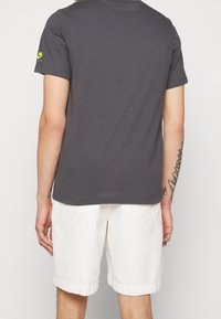 Grå bomullströja med korta ärmar, bakvy. Buren med vita shorts, har en liten grön logotyp på vänster axel.