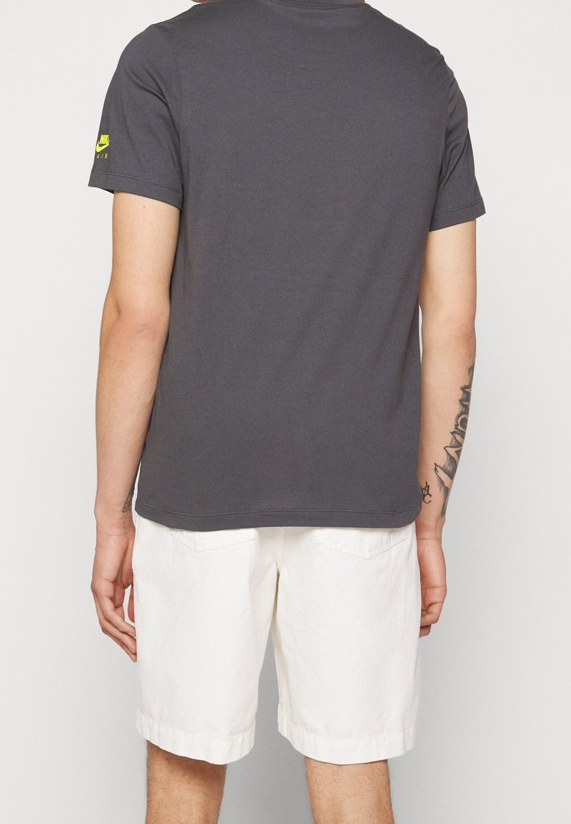 Grå bomullströja med korta ärmar, bakvy. Buren med vita shorts, har en liten grön logotyp på vänster axel.