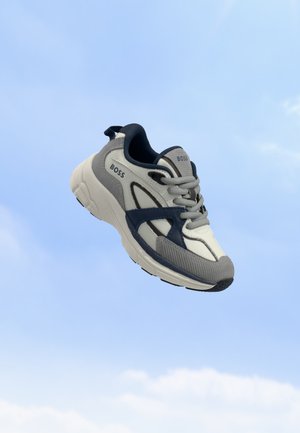 Baskets BOSS blanches et grises avec des accents bleu marine flottant sur un fond de ciel bleu clair.