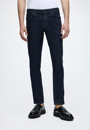 Homme portant un jean slim bleu foncé, des mocassins en cuir noir et une chemise blanche, debout avec les bras détendus le long du corps.