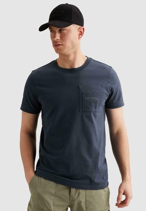 Mężczyzna w czarnej czapce i granatowym krótkim rękawie z kieszenią z logo "PME Legend", w połączeniu z oliwkowozielonymi szortami cargo, ręce w kieszeniach.