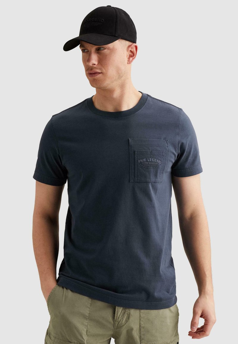 Homme portant une casquette noire et un t-shirt bleu marine à manches courtes avec une poche et le logo "PME Legend", associé à un short cargo vert olive, les mains dans les poches.