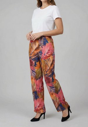 Donna in piedi che indossa una maglietta bianca a maniche corte, pantaloni larghi a stampa tropicale colorata e scarpe nere con tacco su sfondo grigio chiaro.