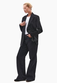 Nainen, jolla on vaaleat hiukset nutturalla, pukeutuu tummansiniseen bleiseriin, jossa on ribattuja hihoja, valkoiseen paitaan, leveisiin mustiin farkkuihin ja mustiin kenkiin. Hän seisoo käsi taskussa.