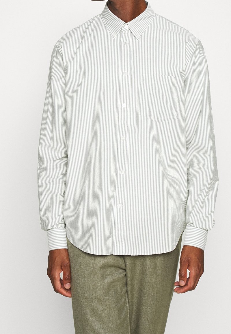 Homme portant une chemise blanche à boutons avec de fines rayures verticales grises et un pantalon vert olive clair, debout devant un fond uni.