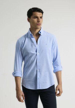 Camisa azul claro de botones, hecha de tejido entrelazado, con cuello y mangas largas enrolladas en los puños. Textura sutil y pequeño logo.