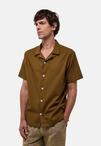 Chemise à manches courtes, à motif marron avec un col boutonné et des boutons blancs, fabriquée en tissu léger avec une finition texturée.