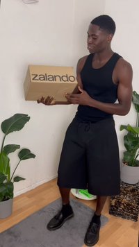 Caixa castanha da Zalando com logótipo preto segurada por uma pessoa, vestindo uma t-shirt preta e calções, em pé sobre um tapete cinza peludo perto de plantas verdes.