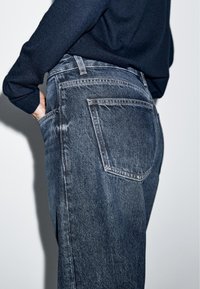 Personne portant un jean bleu foncé avec une poche arrière visible et un haut à manches longues foncé, main glissée dans la poche avant.