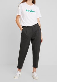 Vit bomullströja med grön "benetton"-logga, kombinerad med grå veckade byxor och vita sneakers. Enkel, avslappnad passform.