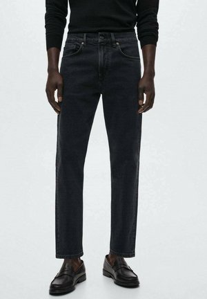 Mężczyzna w czarnych prostych dżinsach, czarnej koszuli z długim rękawem oraz czarnych skórzanych mokasynach na jednolitym szarym tle.