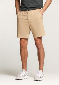 Pantalones cortos beige con frente plano, dos bolsillos laterales y cierre de botón. Cintura plana, tejido de algodón, corte hasta la rodilla. Zapatillas blancas.