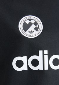 Melnas auduma krekls ar apaļu balto logo, kas satur ģeometrisku rakstu un tekstu, kā arī lieliem baltiem burtiem, kas veido vārdu "adidas".