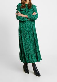 Groene luipaardprint jurk met lange mouwen, ruches, gelaagd ontwerp en een losse pasvorm. Gecombineerd met zwarte enkellaarsjes.