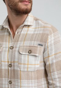 Camicia a quadri marrone e bianca con dettagli gialli, dotata di colletto button-down e tasche sul petto. Realizzata in tessuto testurizzato con bottoni in legno.