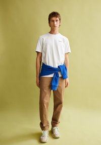 T-shirt blanc à manches courtes, sweatshirt bleu noué à la taille, pantalon beige et baskets claires sur un fond vert uni.