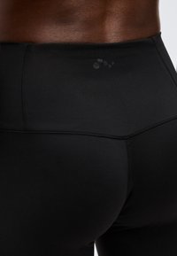 Κοντινή λήψη ατόμου που φοράει μαύρα αθλητικά κολάν ψηλής μέσης με μικρό κυκλικό λογότυπο στη ζώνη, δείχνοντας την πλάτη και την περιοχή της μέσης.