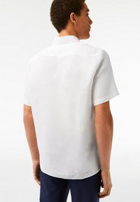 Camicia bianca a maniche corte realizzata in un tessuto leggero, con una texture liscia e un design con colletto classico, vista da dietro.