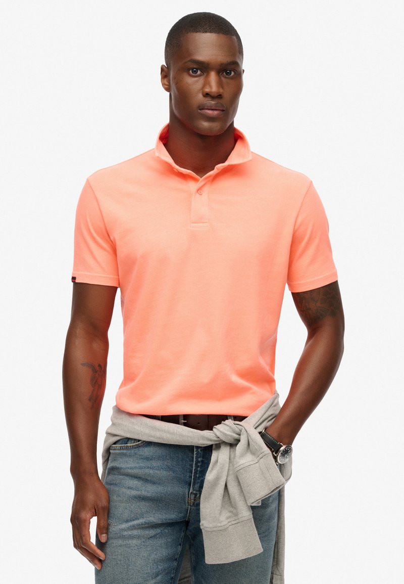 Koraalkleurig katoenen polo shirt met een kraag en twee knopen, gecombineerd met blauwe jeans en een grijze sweatshirt geknoopt om de taille. Horloge om de pols.