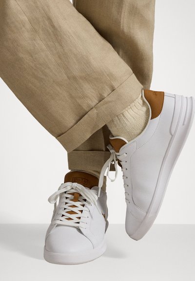 Hvite sneakers med brune detaljer, med en glatt lærtekstur, flate snøringer og en logo på tunga, parret med beige linbukser.