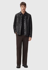 Zwarte leren jas met een knoopsluiting aan de voorkant, vier voorzakken en een kraag, gecombineerd met donkerbruine broek.