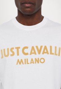 Bílý bavlněný tričko s kulatým výstřihem s výrazným žlutým nápisem: "JUST CAVALLI MILANO." Hladká textura, ležérní design.