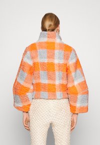 Mønstret cropped jakke i orange, grå og hvid tern. Har en høj krave og tekstureret stof med puffede, brede ærmer.