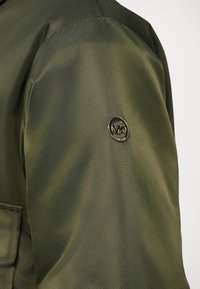 Oliivroheline jakk, millel on sile ja veidi läikiv tekstuur. Vasakul varrukal on ümmargune metallist logo silt.