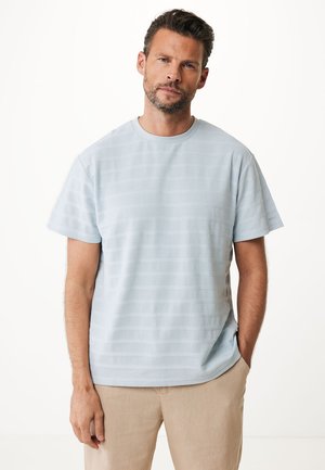 Lichtblauw t-shirt met korte mouwen en textuur met horizontale strepen. Ronde halslijn, relaxte pasvorm en zachte stof. G gedragen met beige broek.