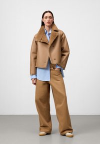 Braune kurz geschnittene Jacke mit einem Lammfellkragen, kombiniert mit einem hellblauen Hemd und weiten beige Hosen. Das Schuhwerk ist hell gefärbt mit einem grünen Akzent.