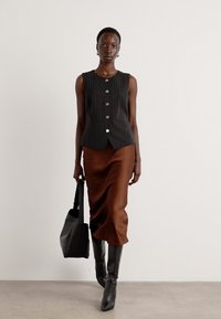 Top senza maniche a righe nere con bottoni, abbinato a una gonna in satin marrone. La modella tiene una borsa a tracolla nera, indossando stivali neri al ginocchio.