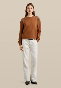 Sudadera marrón con decoración de cuentas en los hombros, combinada con pantalones blancos de pierna ancha y zapatos de tacón negros. Textura suave, corte informal.