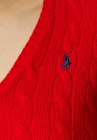 Pull rouge en maille torsadée avec un col en V profond et un petit logo polo bleu marine brodé sur la poitrine. Tissu en laine douce et texturée.