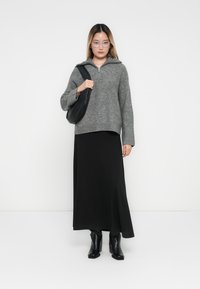 Grijze trui met ritssluiting bij de hals en wijde mouwen, gecombineerd met een zwarte maxi-rok en zwarte enkellaarsjes. Model draagt een zwarte schoudertas.