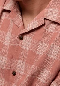 Camisa de botones de manga corta, de color rosa claro, con un patrón de cuadrícula. La tela tiene un acabado texturizado, con botones marrones y un cuello.