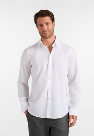 Uomo che indossa una camicia bianca a maniche lunghe abbottonata e pantaloni grigi, sorridente con una mano in tasca, su uno sfondo bianco semplice.