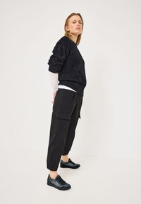 Pullover nero in pelliccia sintetica con spalle scese, abbinato a pantaloni cargo neri con tasche laterali. Scarpe da ginnastica nere completano l'outfit.