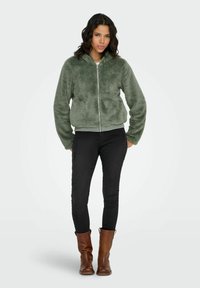 Zielona, pluszowa bluza na zamek z kapturem, o miękkiej fakturze i elastycznych mankietach, zestawiona z czarnymi dopasowanymi spodniami oraz brązowymi kozakami za kolano.