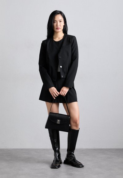 Black cropped jacket with two buttons, matching pleated mini skirt, knee-high black glossy boots, and a structured black handbag.