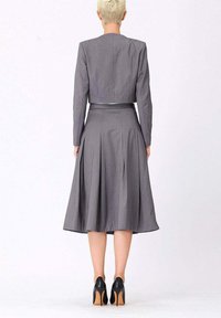 Femme avec des cheveux blonds courts portant une veste cropped à manches longues grise et une jupe midi assortie avec des talons hauts noirs, se tenant de dos.